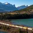 Freight train traveling along turquoise lake in mountain valley with snow-capped peaks and evergreen forest