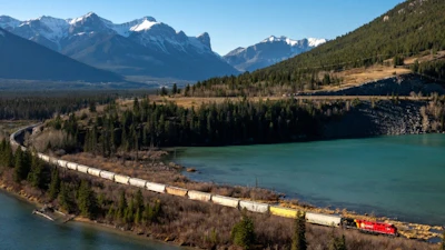 Freight train traveling along turquoise lake in mountain valley with snow-capped peaks and evergreen forest