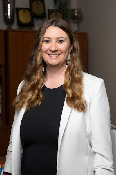 Professional woman with long brown hair wearing white blazer and black top, smiling in office setting with awards in background