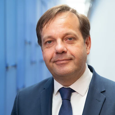 Professional headshot of a man in dark business suit and navy tie against blue office background