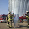 Firefighters in yellow gear conduct training exercises near a large metal grain silo with fire trucks parked in the background.