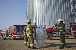 Firefighters in yellow gear conduct training exercises near a large metal grain silo with fire trucks parked in the background.