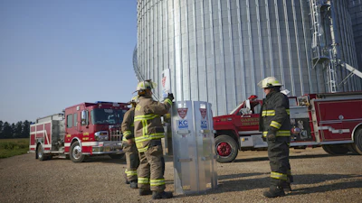 Firefighters in yellow gear conduct training exercises near a large metal grain silo with fire trucks parked in the background.