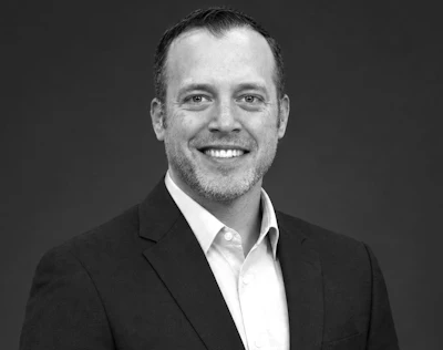 Professional headshot of smiling man in black blazer and white shirt against gray background