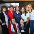 Seven women smiling together at an indoor event, some wearing USA team apparel and medals