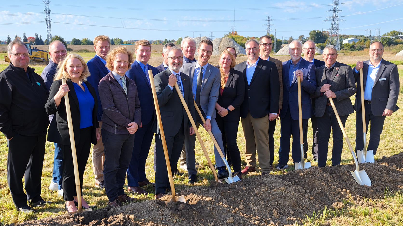 Group of professionals holding shovels at groundbreaking ceremony in rural field with transmission towers in background