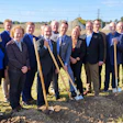 Group of professionals holding shovels at groundbreaking ceremony in rural field with transmission towers in background