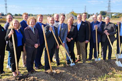 Group of professionals holding shovels at groundbreaking ceremony in rural field with transmission towers in background