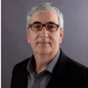 Professional headshot of a man with gray hair wearing glasses and dark blazer over gray shirt