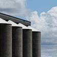 Five cylindrical grain storage silos with metal roof against blue sky and white clouds
