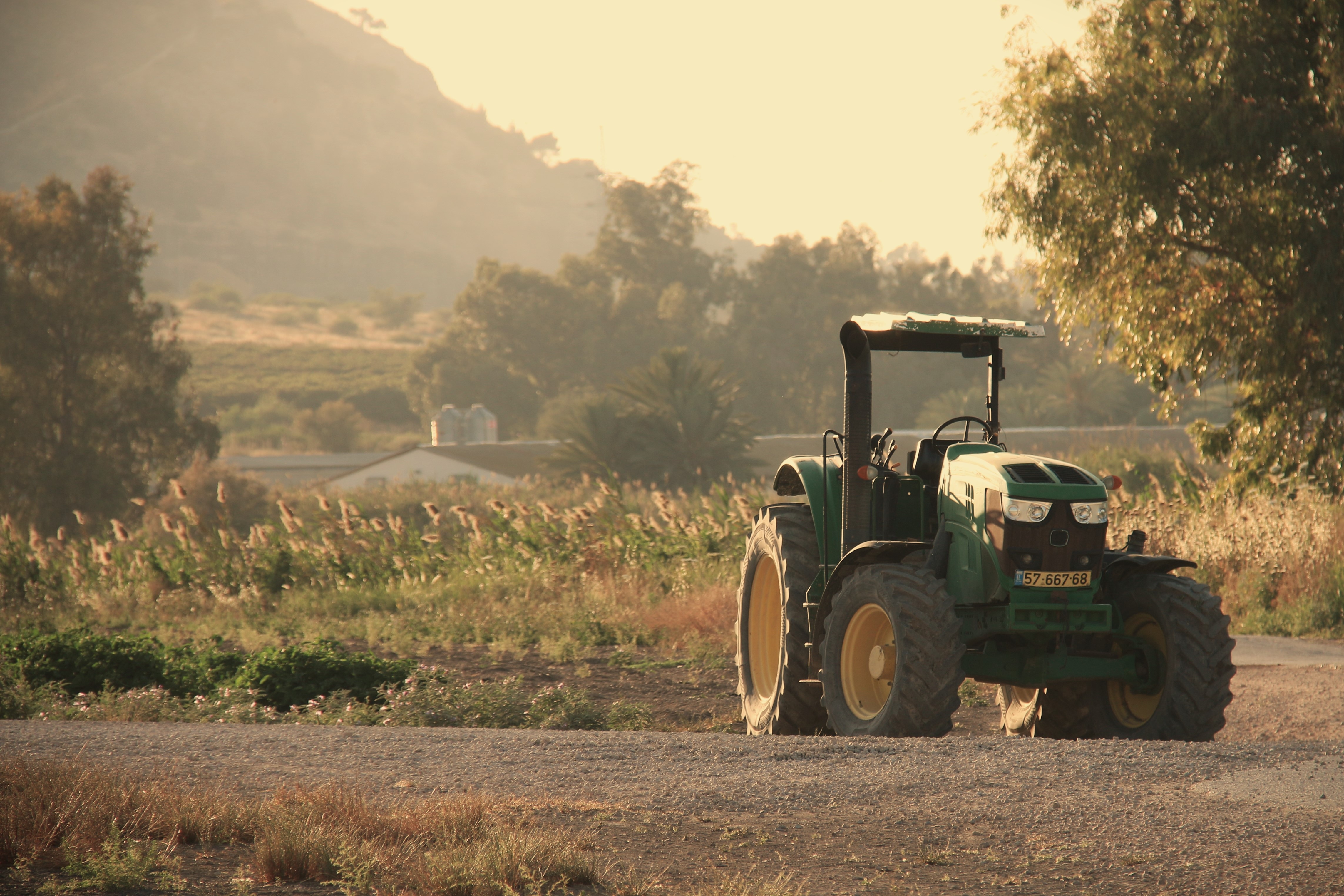 Green tractor parked on rural road with cultivated fields and mountains in misty background at sunset