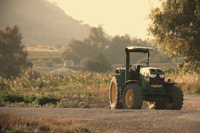 Green tractor parked on rural road with cultivated fields and mountains in misty background at sunset