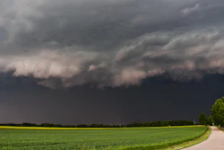 Dramatic storm clouds gather over a green agricultural field with a curved rural road beneath an ominous sky