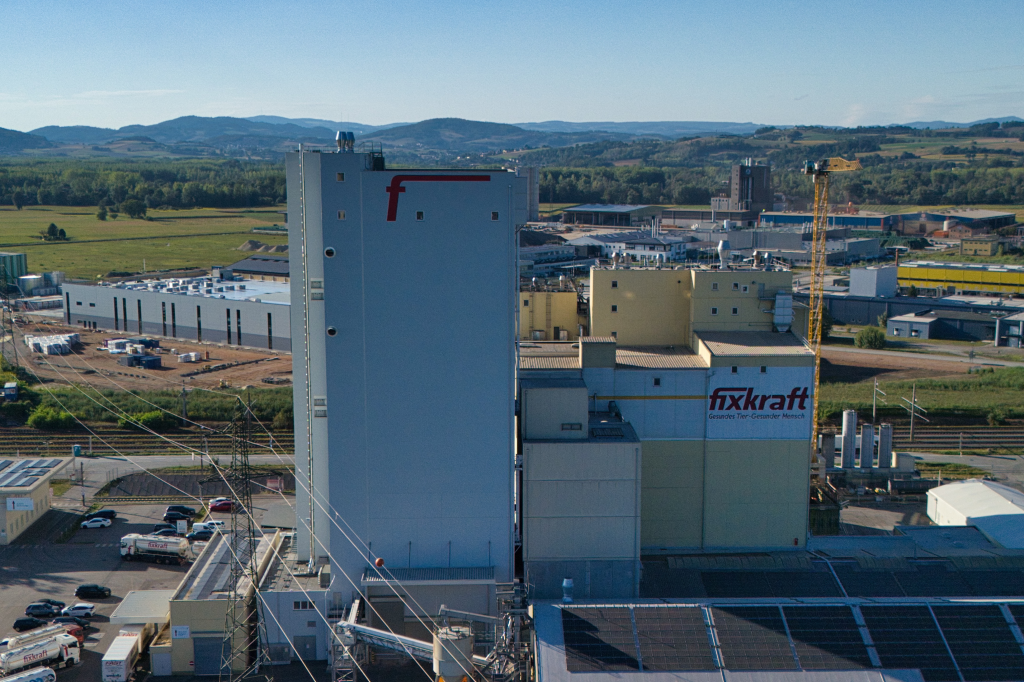 Industrial manufacturing facility with blue silos and fixkraft signage in rural landscape with mountains