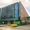 Modern mixed-use building with red brick and glass facade, green architectural accents, and landscaped plaza with yellow ornamental grasses