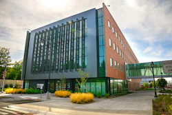 Modern mixed-use building with red brick and glass facade, green architectural accents, and landscaped plaza with yellow ornamental grasses
